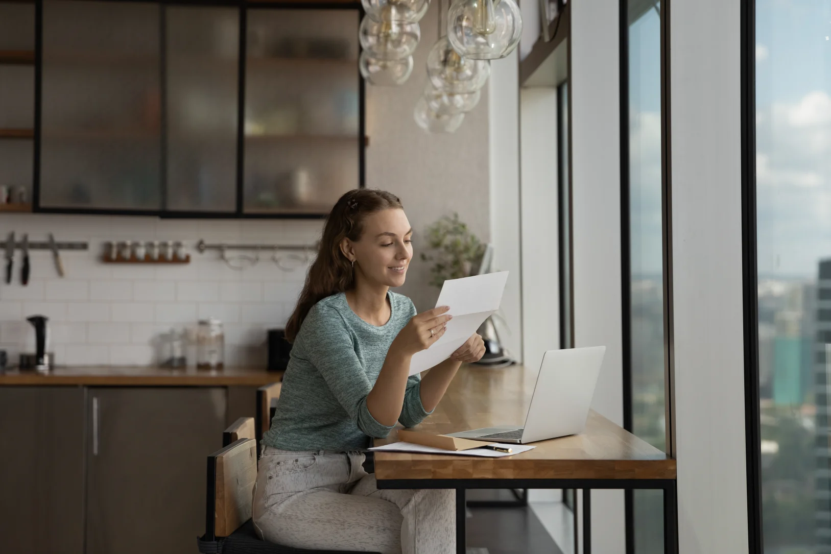 young-woman-reviewing-pay-stub-document-while-working-on-laptop-in-modern-kitchen