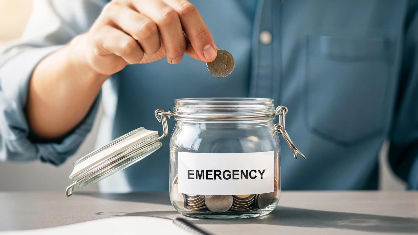 Person placing coin into glass jar labeled emergency fund planning for future Person placing coin into glass jar labeled emergency fund planning for future